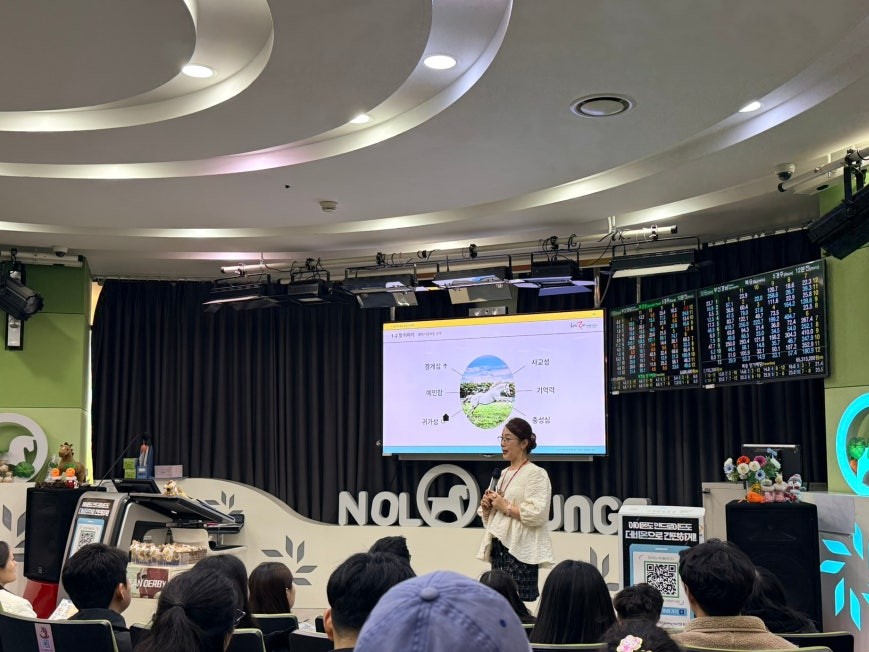 Beginner’s Class session at Let’s Run Park Seoul with a presenter holding a microphone at the front, audience seated in rows, projection screen showing horse racing concepts, and race odds displays on side walls