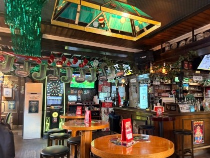 Wide interior view of Rose & Crown with round wooden tables, black stools, a dart board visible in the background, and a green decorative chandelier overhead
