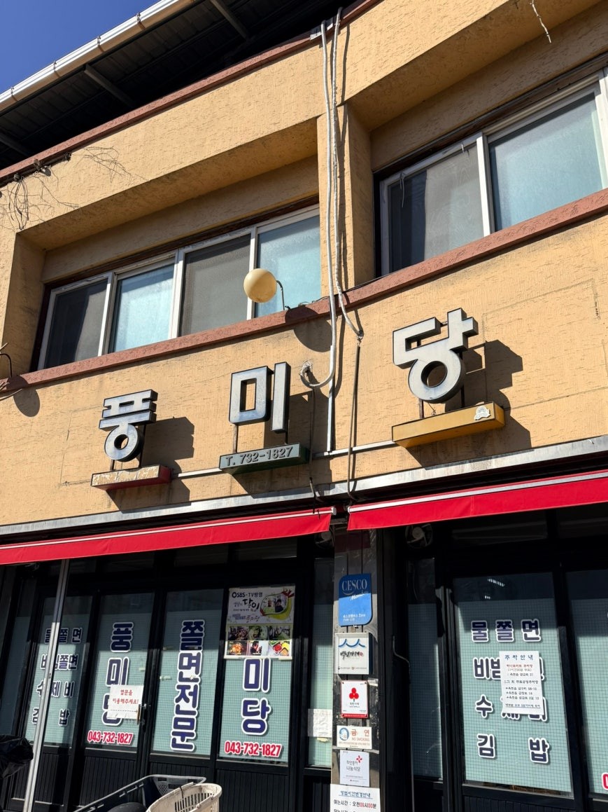 Exterior of Pungmidang restaurant in Okcheon — a weathered yellow two-story building with large bold black lettering spelling 풍미당, a red awning along the ground floor, and menu items including mul-jjolmyeon printed on the windows with an SBS TV appearance sticker near the entrance