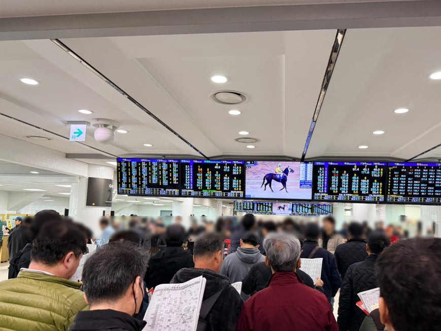 Crowded second floor interior at Let’s Run Park Seoul with multiple large digital odds boards spanning the ceiling, visitors standing and holding race programs, and a center screen showing a racing horse