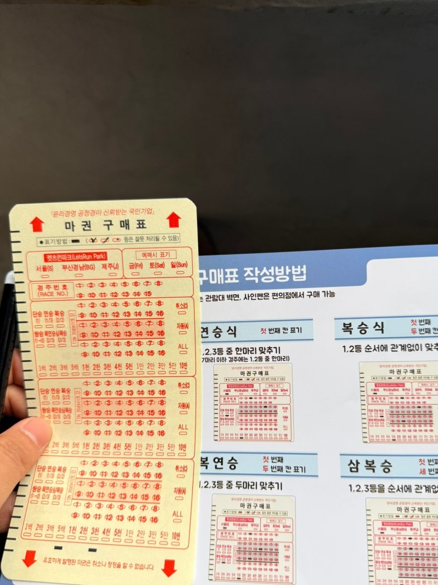 OMR betting card for Let’s Run Park Seoul held in hand showing bubble-fill sections for race and horse numbers, with an instructional poster behind showing betting type categories