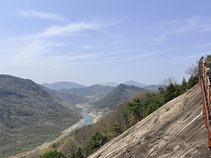 Panoramic view from approximately 80% up Yonggwolsan Sky Trail, showing the Seomjin River curving through a wide mountain valley under clear blue sky, with wooden deck stairs running along the granite cliff face on the right side of the frame
