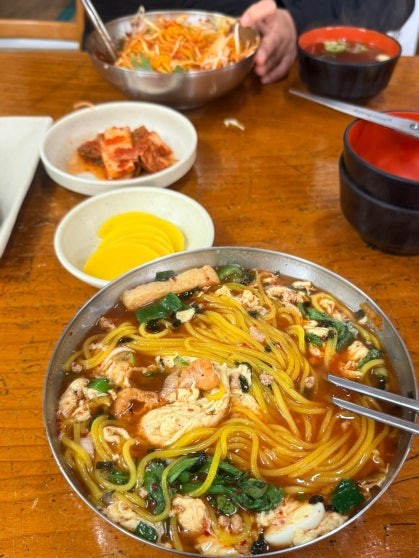 Large metal bowl of mul-jjolmyeon at Pungmidang — thick springy jjolmyeon noodles in a reddish, tangy and lightly spicy cold broth, topped with vegetables and other ingredients, with kimbap rolls and side dishes visible in the background