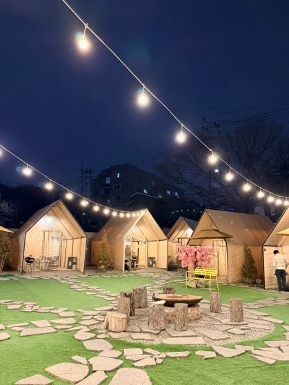 Nighttime view of the outdoor glamping BBQ area at Dosim Sok Grill with lit A-frame tent cabins, stepping stone paths, artificial grass, string lights overhead, and a central log seating area