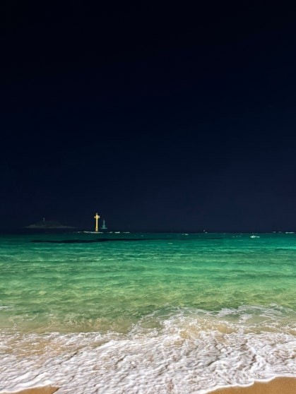 Sokcho Beach (속초해수욕장) at night with luminous emerald-green shallow water catching artificial light, gentle waves rolling onto pale sandy shore, a lighthouse structure visible on the breakwater in the distance