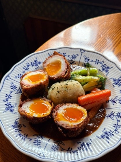Scotch egg halved to show a soft jammy yolk wrapped in seasoned minced meat on a blue-and-white patterned plate with steamed broccoli, carrots, potato mash, and brown gravy