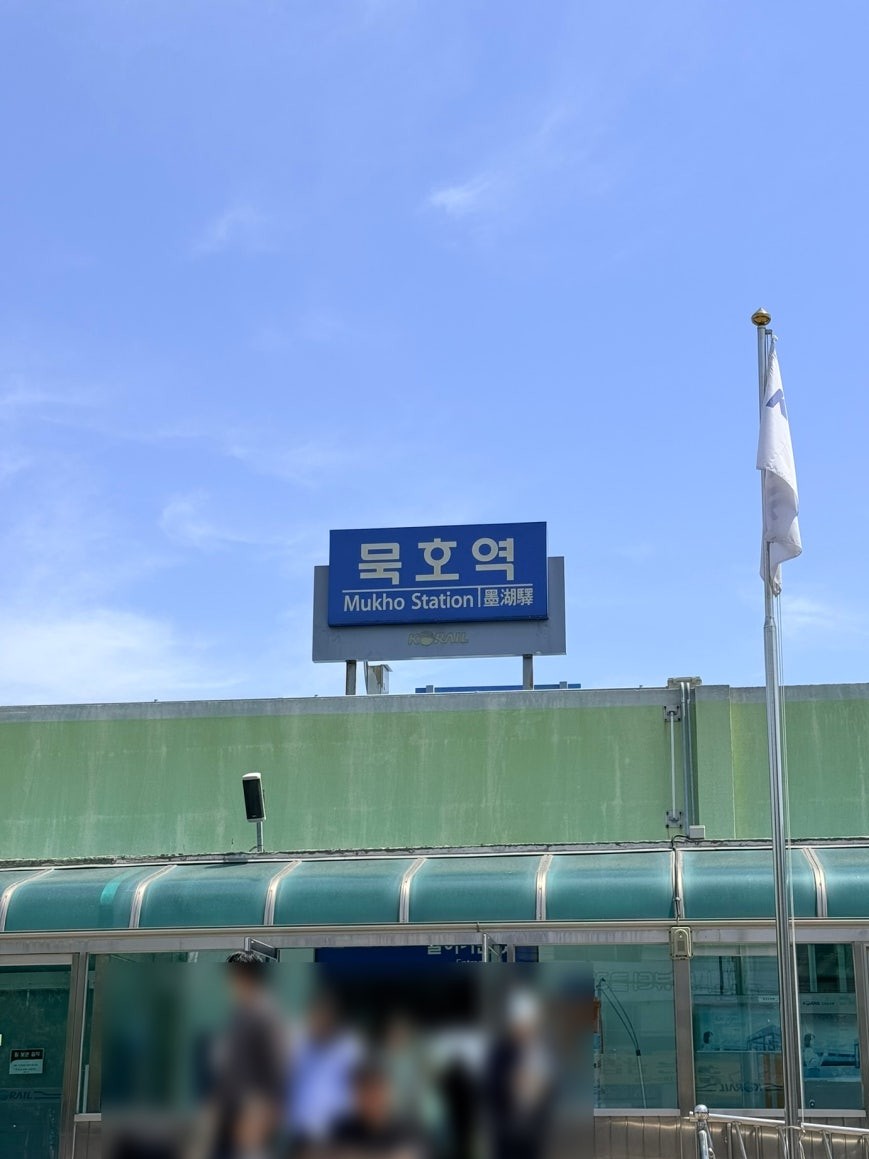 Mukho Station exterior with a large blue bilingual sign reading 묵호역 and Mukho Station in Korean, Chinese, and English, against a clear blue sky, with blurred pedestrians and a flagpole in the foreground