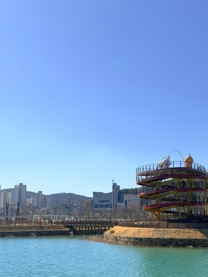Spiral multi-level observation tower in red and yellow rising above the lake at Gapcheon Eco Lake Park, a long suspension bridge stretching across the turquoise water in the middle ground, Daejeon city buildings filling the background under a clear blue sky