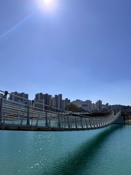Long suspension bridge spanning the turquoise lake at Gapcheon Eco Lake Park, viewed from ground level, with metal railings and sunlight glinting off the water surface and residential towers lining the far shore