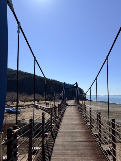 Looking straight down the length of a narrow wooden suspension bridge with steel cable railings and metal support posts, the calm Yellow Sea visible on both sides under a clear blue winter sky