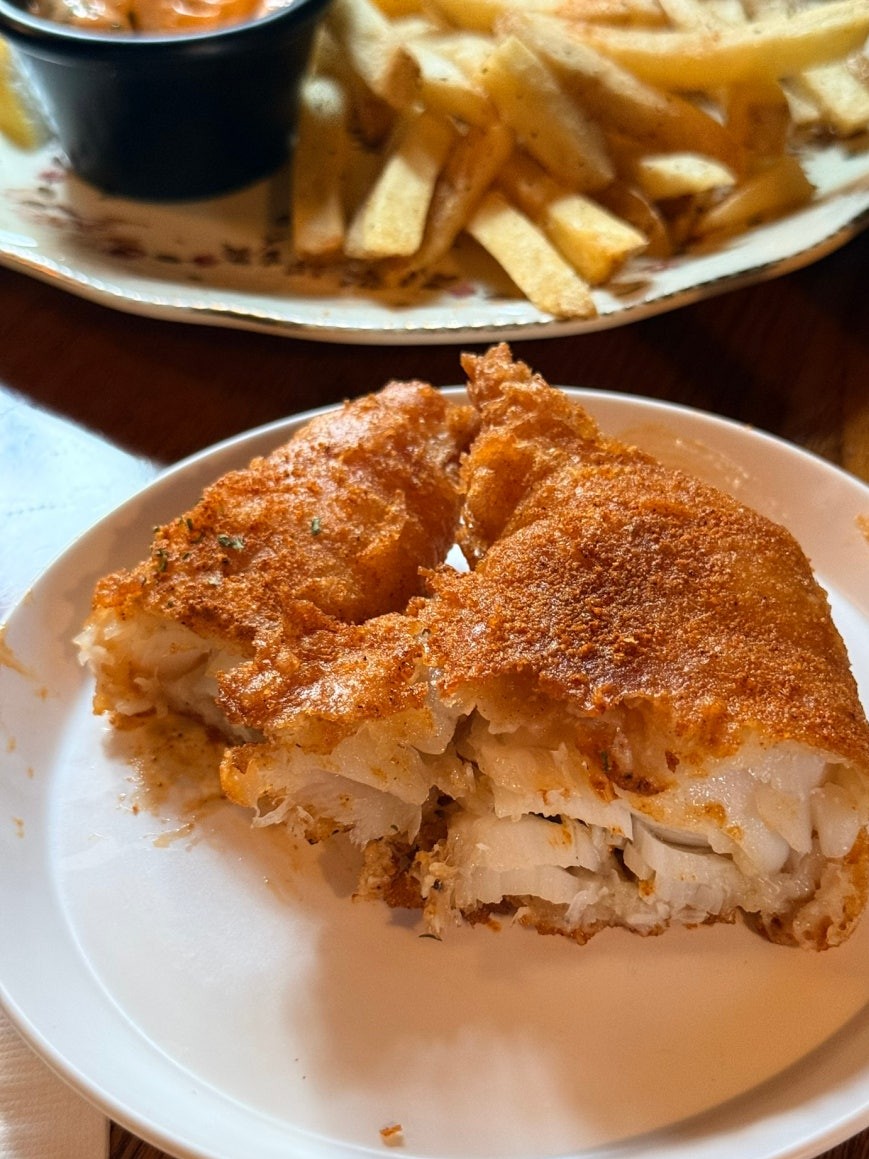 Close-up of fish from Rose & Crown fish and chips, broken open to show thick flaky white flesh inside golden batter, with fries and orange dipping sauce in the background