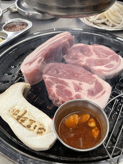 Three thick raw Jeju black pork cuts on a round charcoal tabletop grill with a small metal bowl of spiced dipping sauce, and sliced raw onion visible in the background