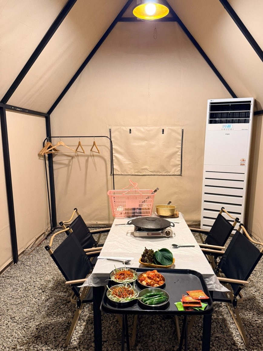 Inside the dining tent at Dosim Sok Grill with a long table covered in white paper, camp chairs on both sides, a cast-iron grill plate on a portable burner, and side dishes including kimchi, greens, and peppers