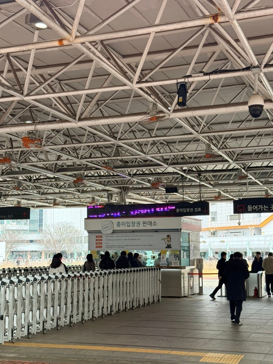 Paper ticket booth at Let’s Run Park Seoul entrance, with folding metal turnstile barriers, visitors queuing at the counter, and a bilingual entrance sign