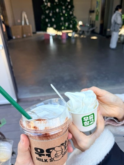 Two hands holding drinks from Boryeong Milk Warehouse Café — a large iced latte in a branded 우유창고 plastic cup and a small soft-serve ice cream cup, with a Christmas tree decorated café entrance visible behind