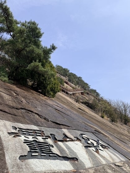 Looking up at Yonggwolsan cliff from below, with large Chinese characters carved into a granite slab in the foreground and the wooden deck trail structure visible high above against a clear blue sky