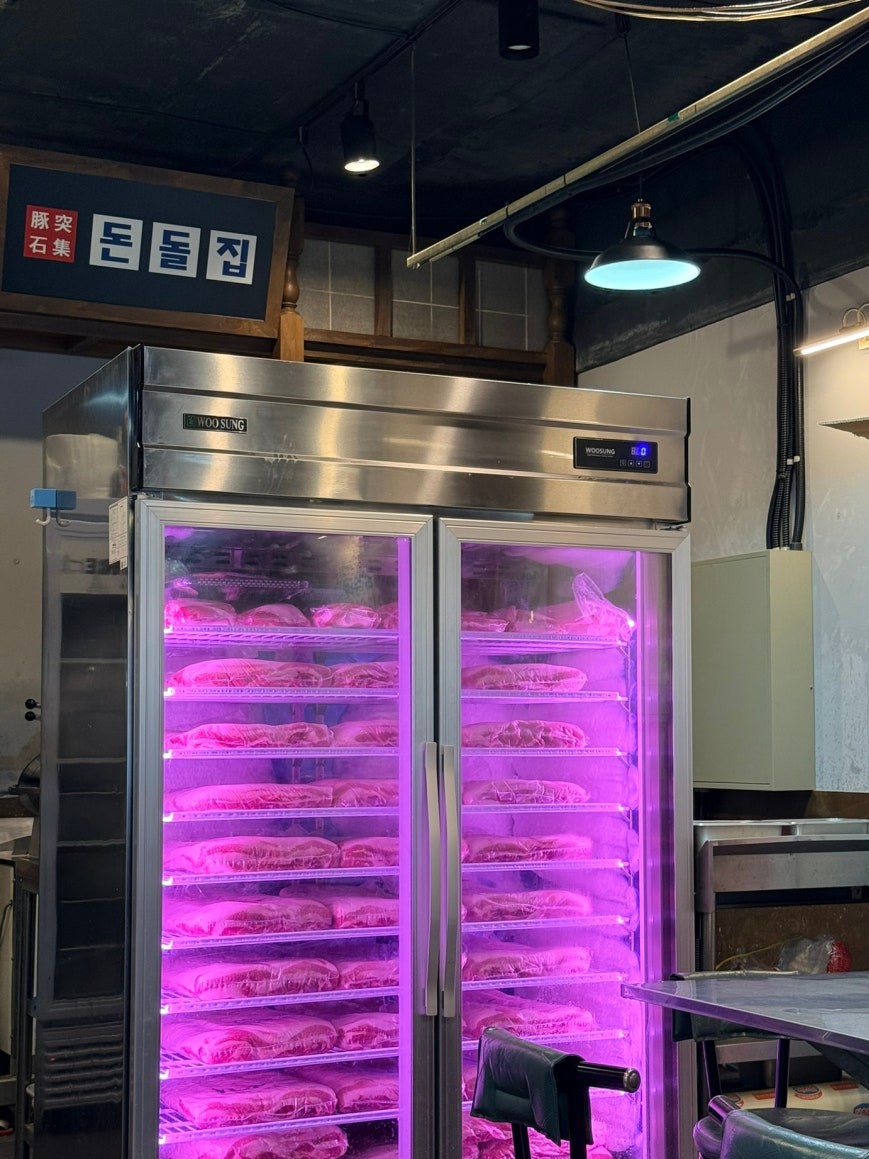 Large stainless steel double-door refrigerator packed with trays of fresh thinly sliced pork belly on multiple shelves, lit with pink interior lighting, with the restaurant sign 돈돌집 visible on the back wall