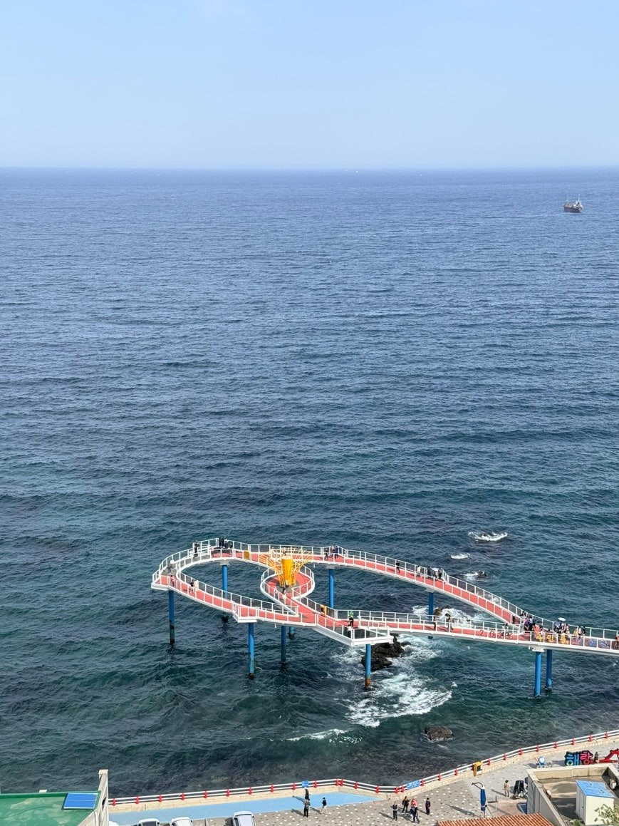 Haerang Observation Deck seen from Dojaebigol Sky Valley above: a curved red-and-white walkway on blue support columns jutting over the East Sea, a fishing vessel visible offshore