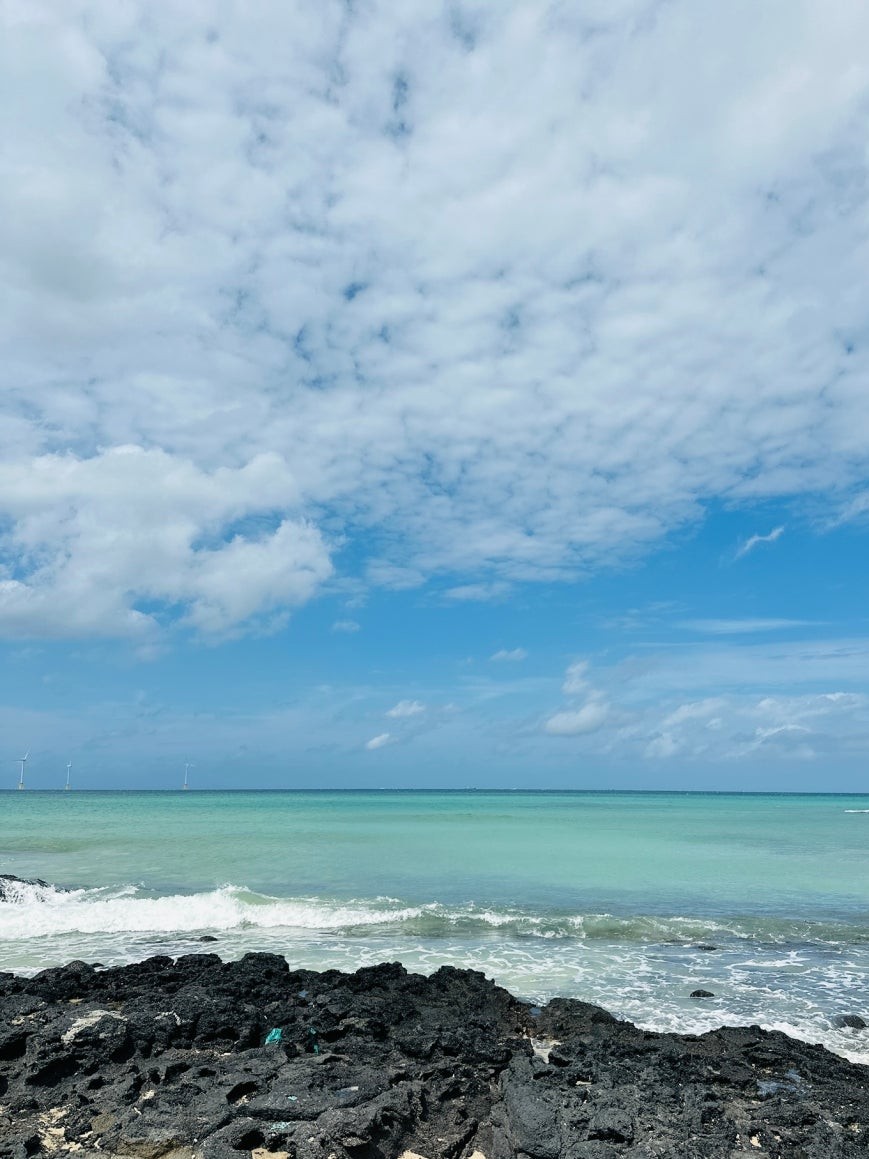 Gwakji Beach in western Jeju with turquoise waves breaking over jagged black volcanic rocks, a strip of pale sand along the shore, and wind turbines on the distant horizon under a partly cloudy sky