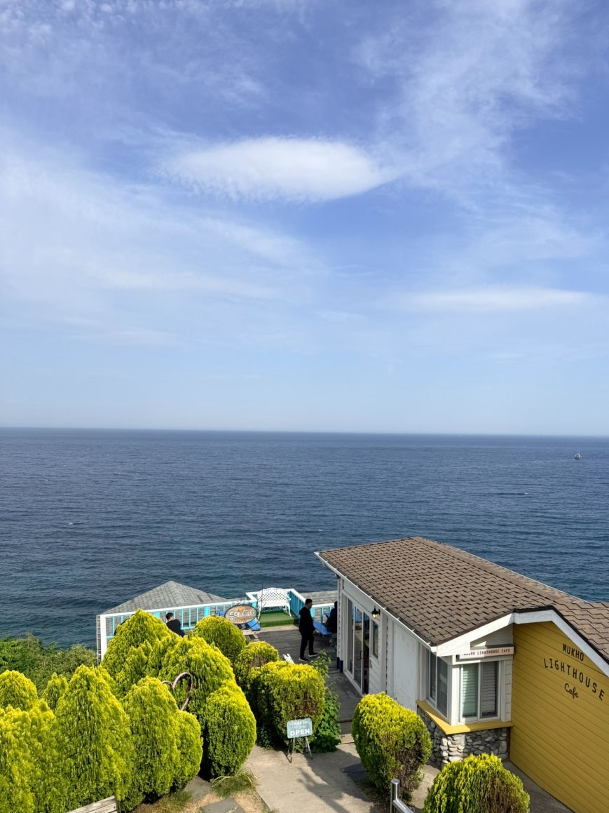 Nongolddam-gil hillside café area: Mukho Lighthouse Cafe with a yellow building, hedge-lined stone path, and a small white terrace overlooking the deep blue East Sea stretching to the horizon