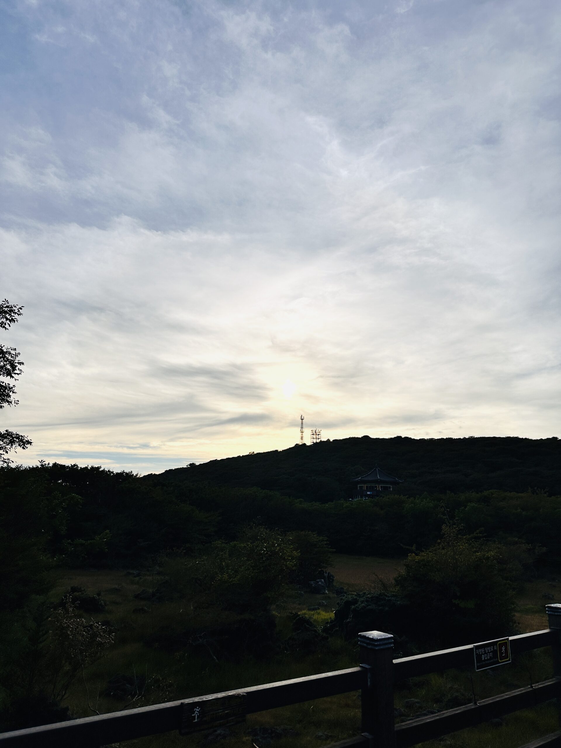 Wooded hillside in Seogwipo with a wooden rail fence in the foreground, a traditional Korean-roofed pavilion visible among the dense trees on the slope and antenna towers at the ridgeline under a partly cloudy sky