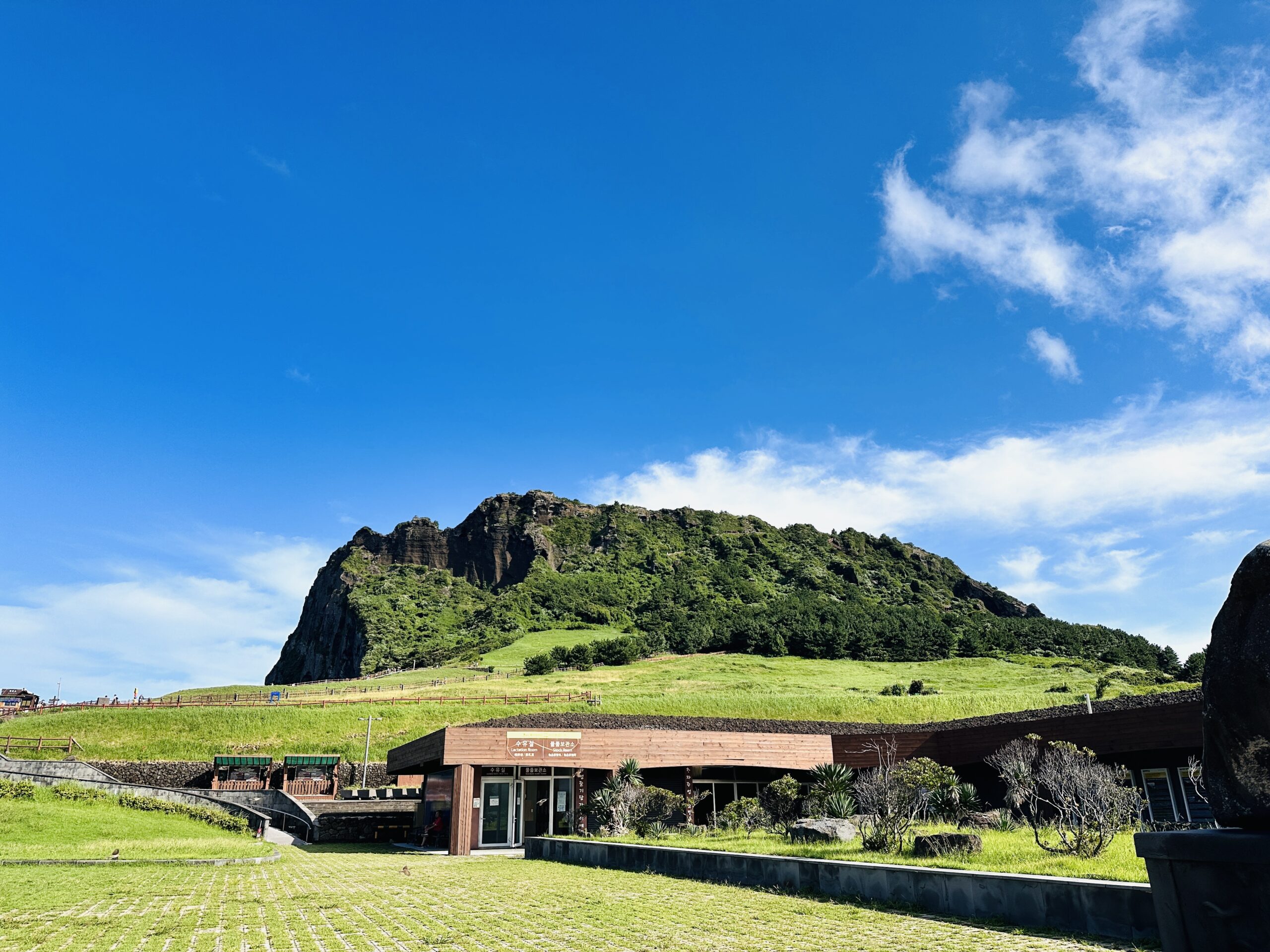 Low wooden-paneled visitor center building nestled against the grassy base of Seongsan Ilchulbong (성산일출봉), the craggy volcanic summit rising above green slopes under a clear summer sky