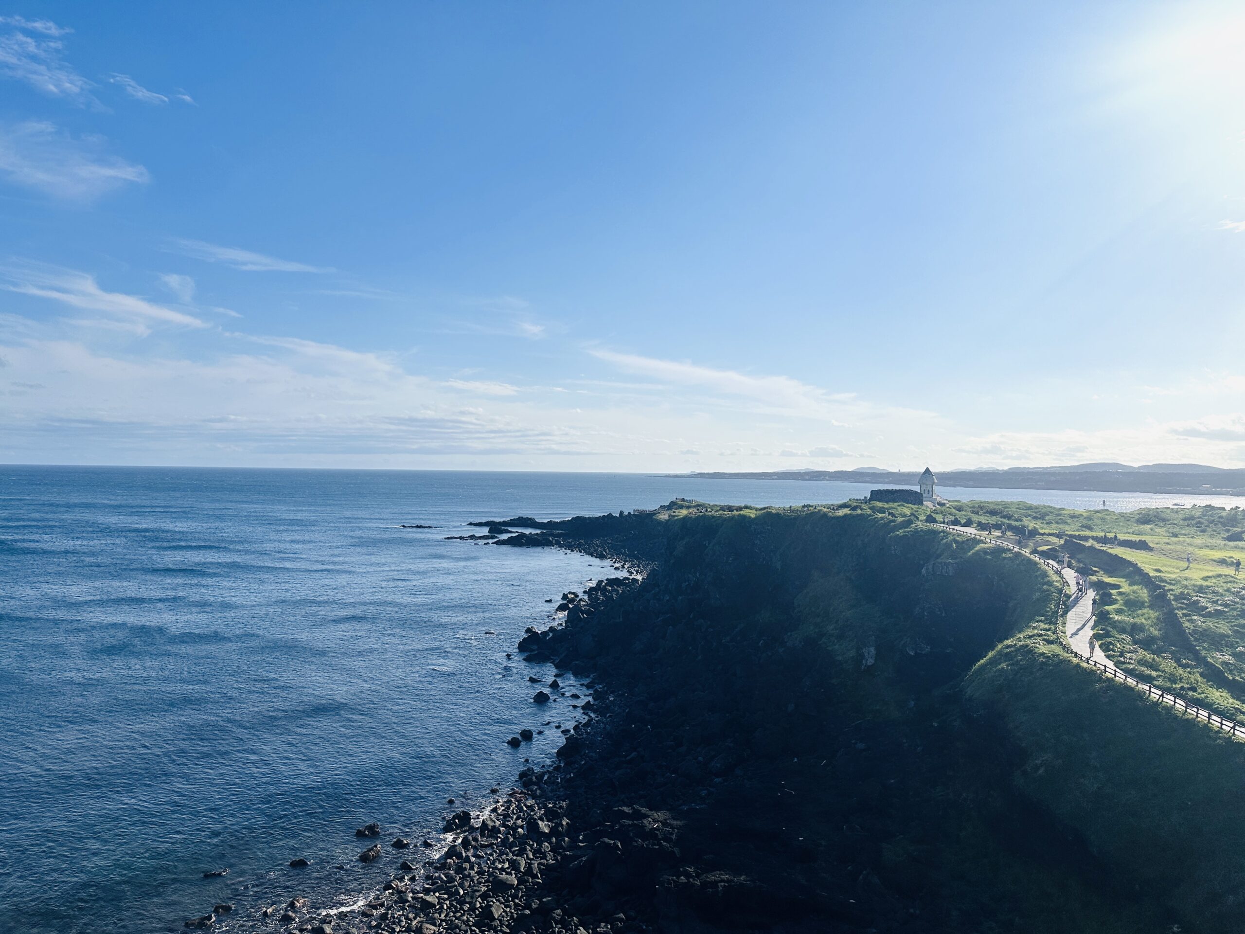 Elevated view of a green coastal headland on Jeju’s south coast curving toward a small white lighthouse-like structure at the point, a narrow walking path with wooden railings following the cliff edge above dark rocks and calm blue sea