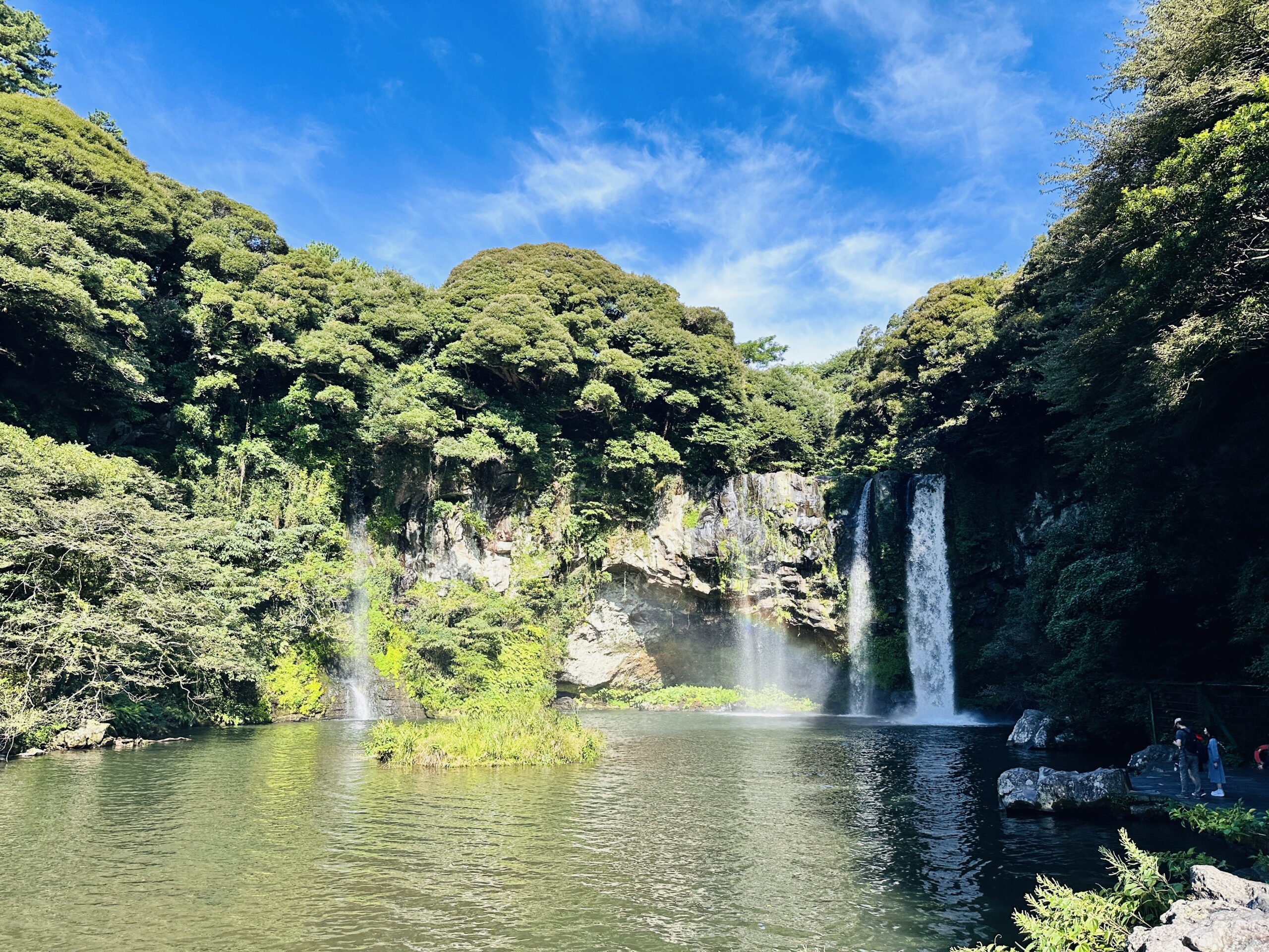 Two waterfalls — one wide curtain on the right, one narrower plume on the left — cascading into a broad emerald natural pool at Cheonjiyeon (천지연폭포), Seogwipo, a small forested island at the pool’s center, dense green trees framing the scene under blue sky