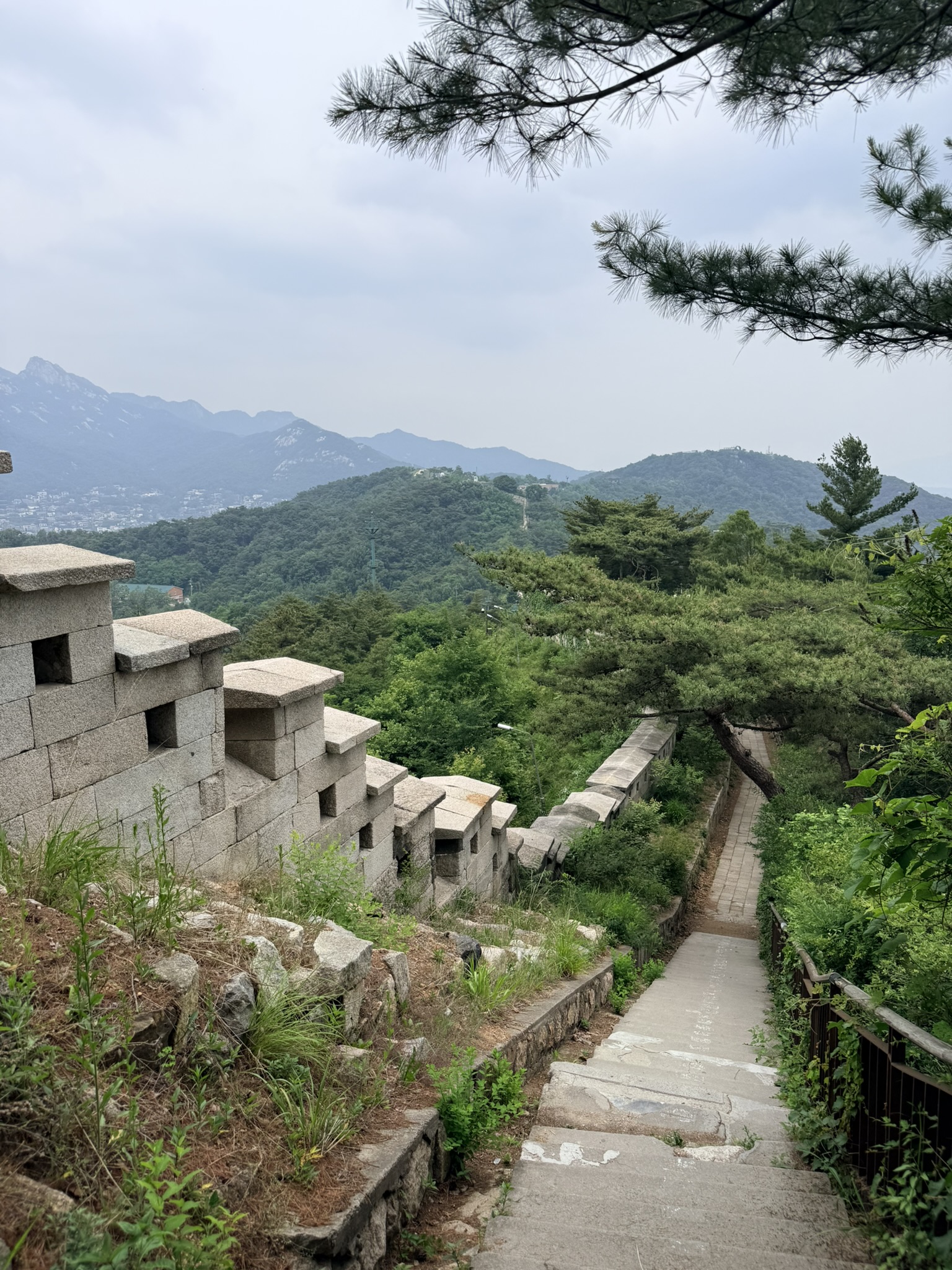 Hanyangdoseong granite battlements with crenellated merlons and arrow slits descending steeply along the Bugaksan ridgeline, stone trail steps with metal railing on the right, Bukhansan mountain range with granite peaks in the background