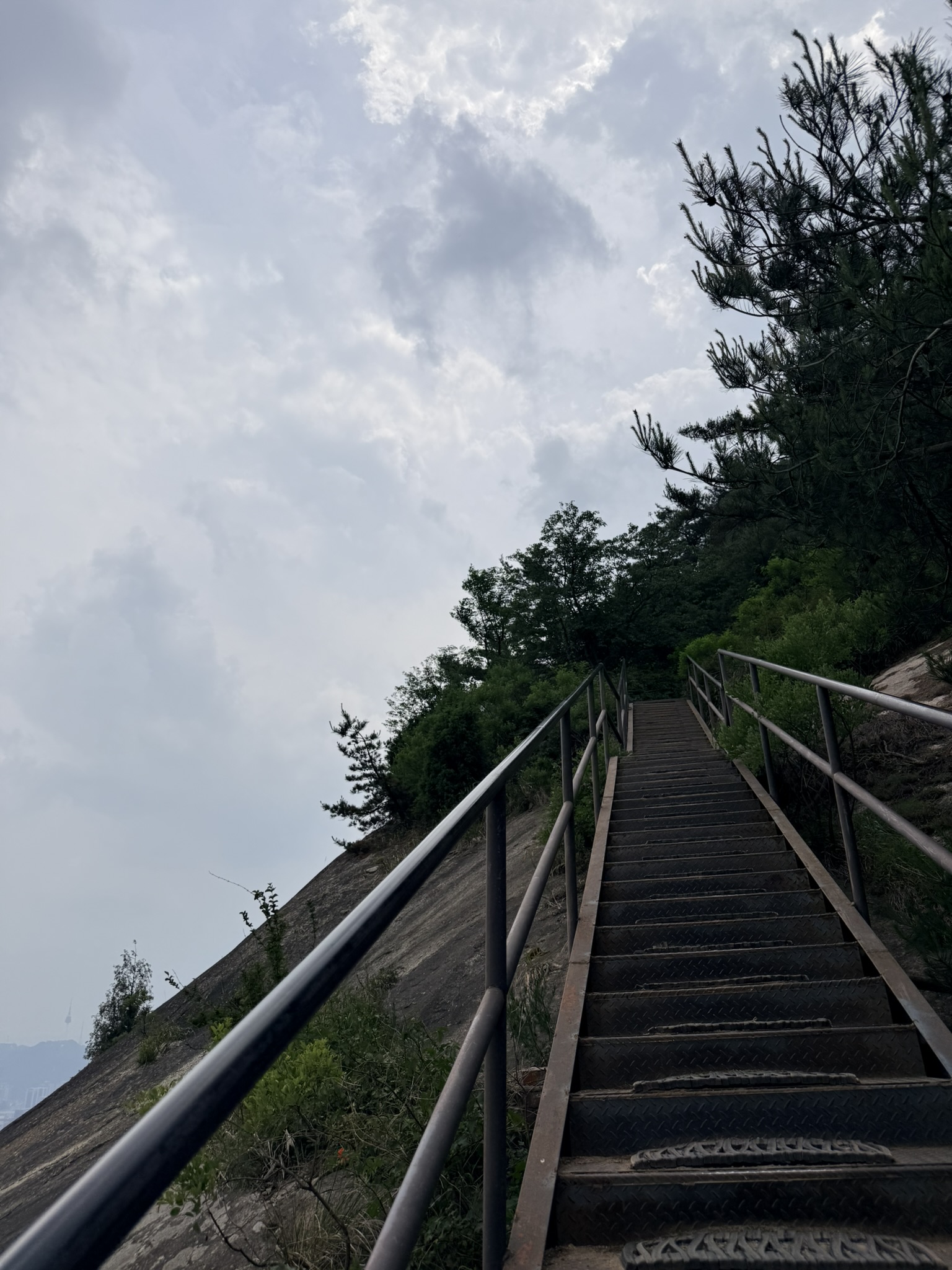 Steep metal staircase ascending bare granite rock face on the final approach to Inwangsan summit, pine trees silhouetted against storm clouds, viewed from below looking upward